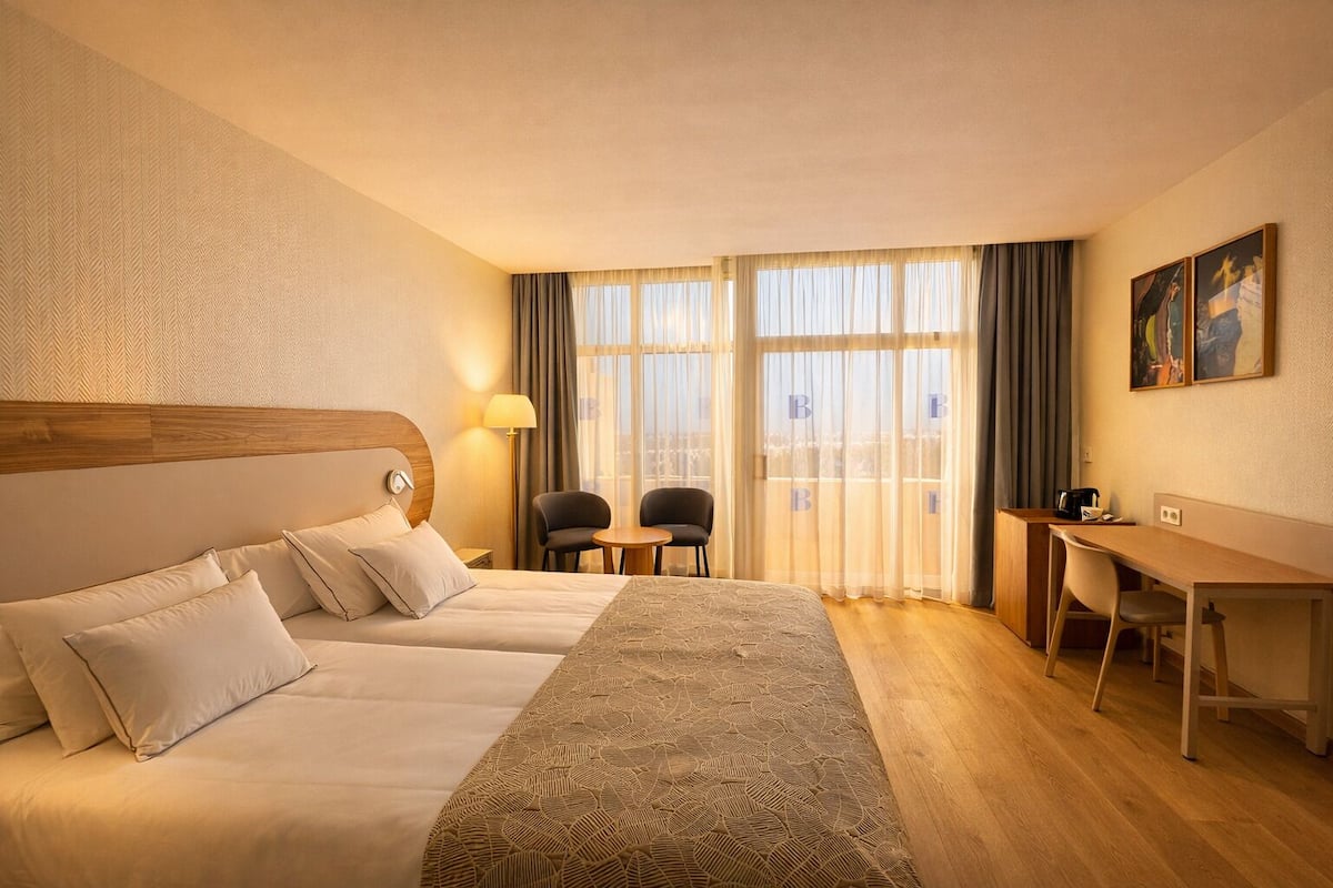 A hotel room with two beds, a desk, chairs, a lamp, and large windows with curtains.