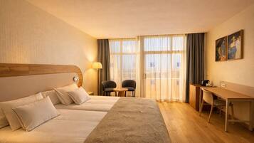 A hotel room with two beds, a desk, chairs, a lamp, and large windows with curtains.