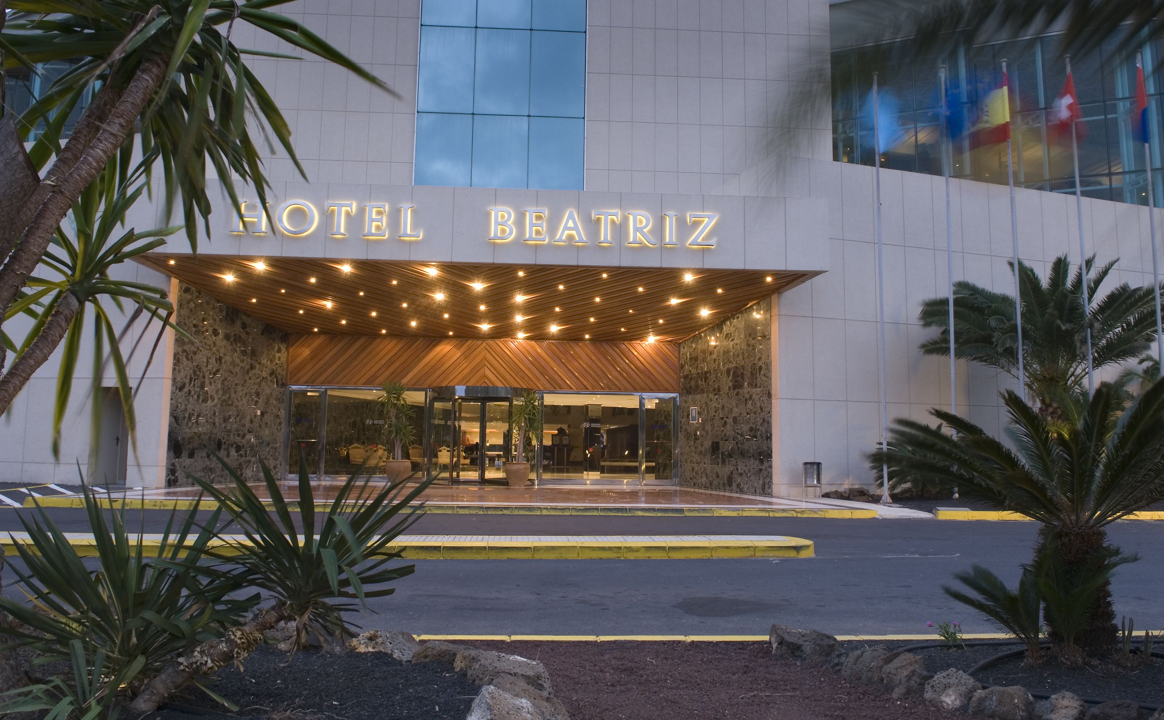 La entrada del Hotel Beatriz, con fachada moderna, amplios ventanales de vidrio y un recibidor acogedor con palmeras y un toldo de madera.