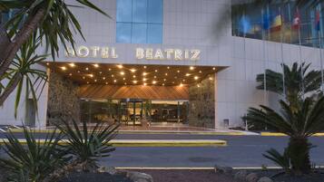La entrada del Hotel Beatriz, con fachada moderna, amplios ventanales de vidrio y un recibidor acogedor con palmeras y un toldo de madera.