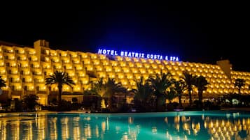 A large hotel building with a pool in front, illuminated by warm lights.