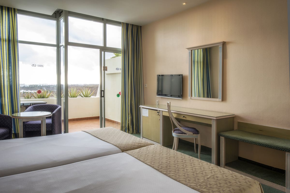 A hotel room with two beds, a desk, a chair, a TV, and a balcony with a view.