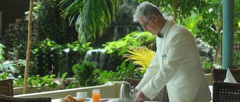 A man in a white jacket preparing coffee at an outdoor table with a view of a waterfall and lush greenery.