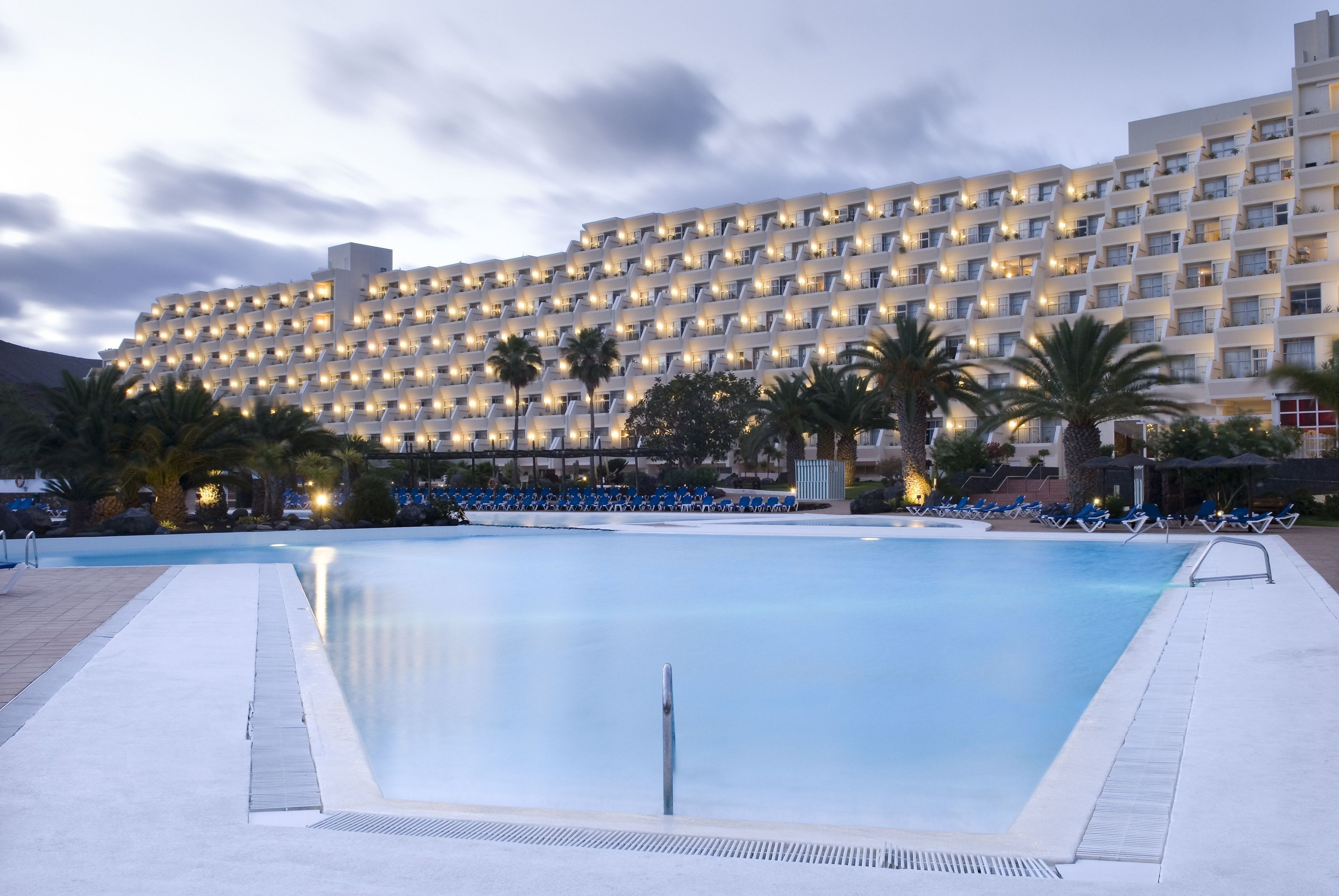 A hotel with a large swimming pool, multiple balconies, and palm trees.