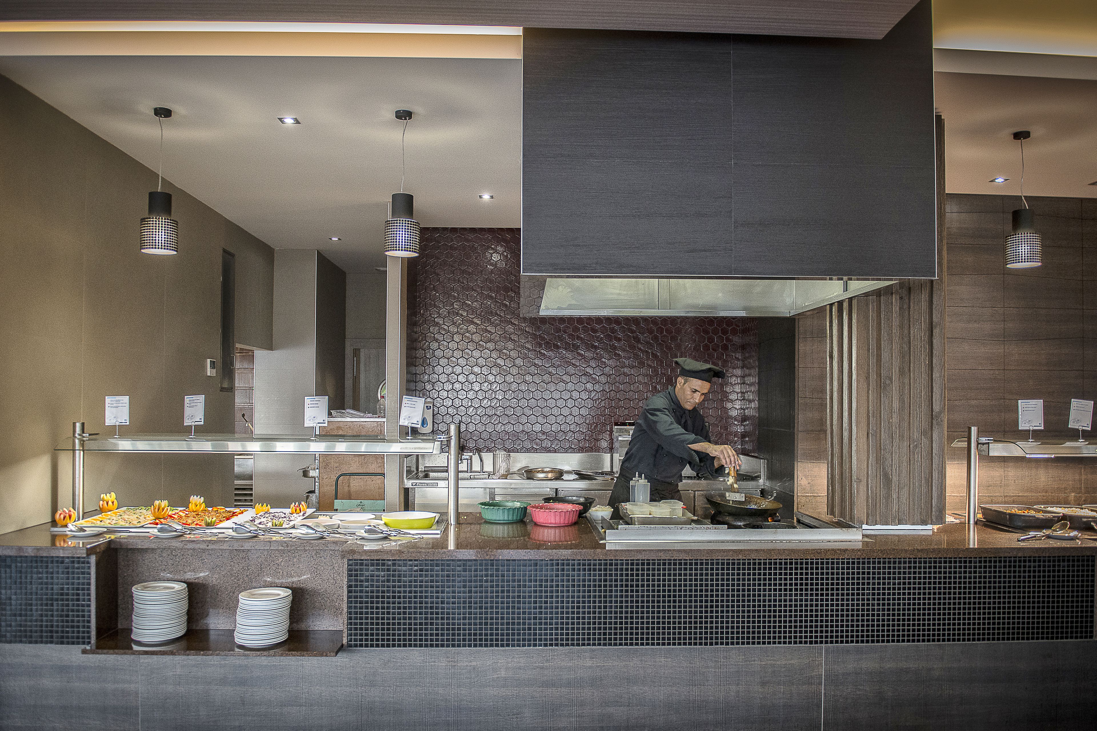 A modern kitchen with a chef preparing food, a buffet with various dishes, and a clean, contemporary design.