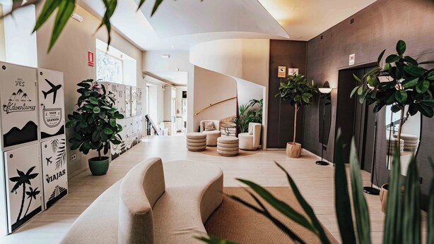 Un moderno vestíbulo de hotel con un sofá curvo, plantas en macetas y una pared con ilustraciones de temática viajera.