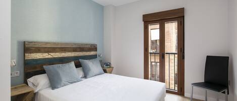Un dormitorio con cabecera de madera, cama con ropa de cama blanca, mesita de noche, silla y ventana con vista a un edificio.