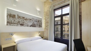 Habitación de hotel con una cama grande, una mesita de noche, una lámpara de pared, una ventana con cortinas y un cuadro de un paisaje urbano.