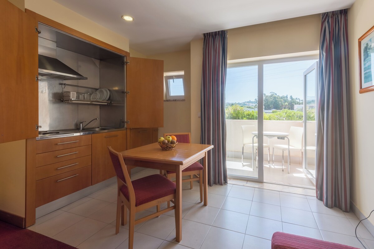 A compact kitchen with wooden cabinets, a dining table with two chairs, and a balcony with a view.