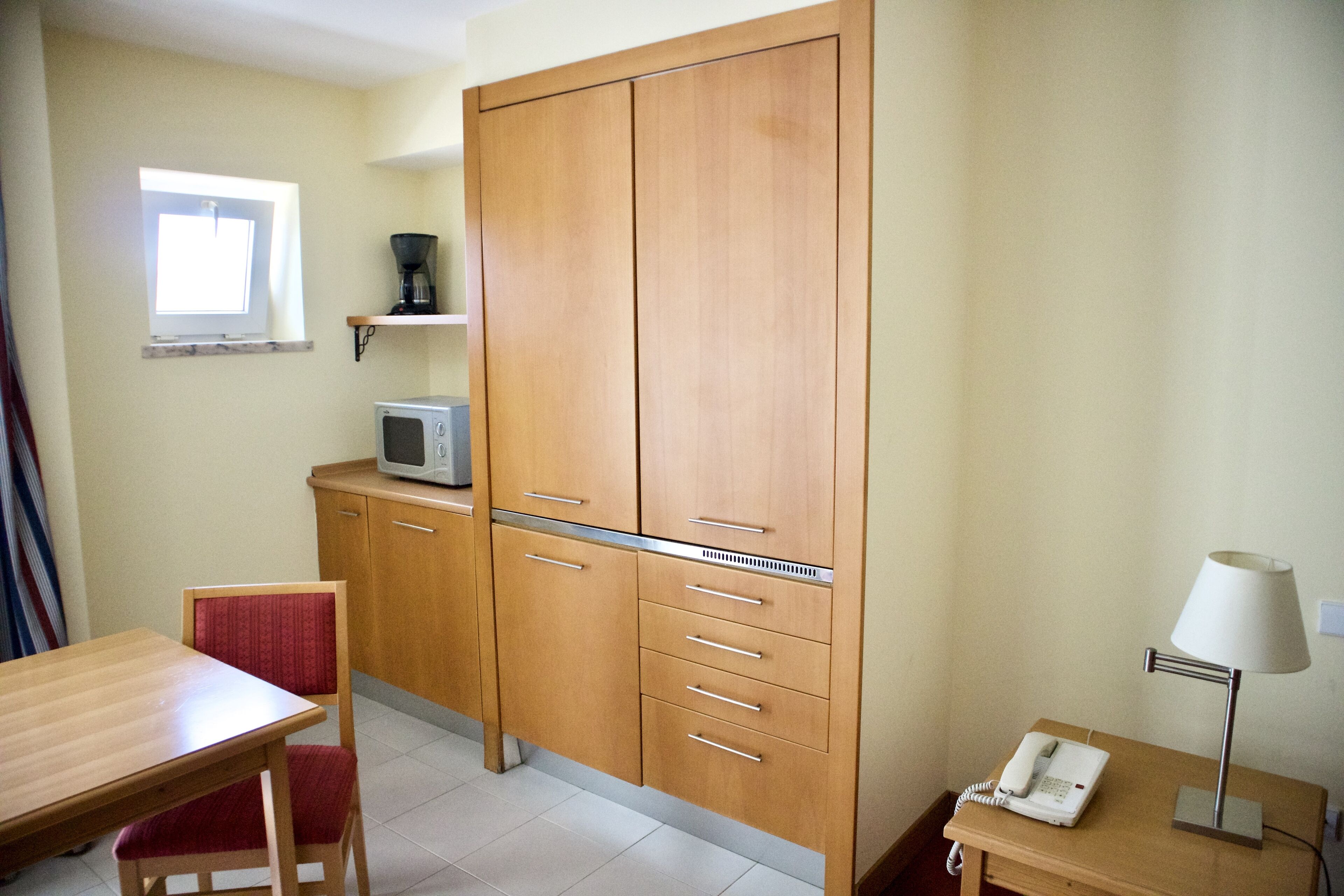 Ein kleines Zimmer, ausgestattet mit einem Holzschrank, einer Mikrowelle, einem Telefon und einem kleinen Tisch mit Stuhl.