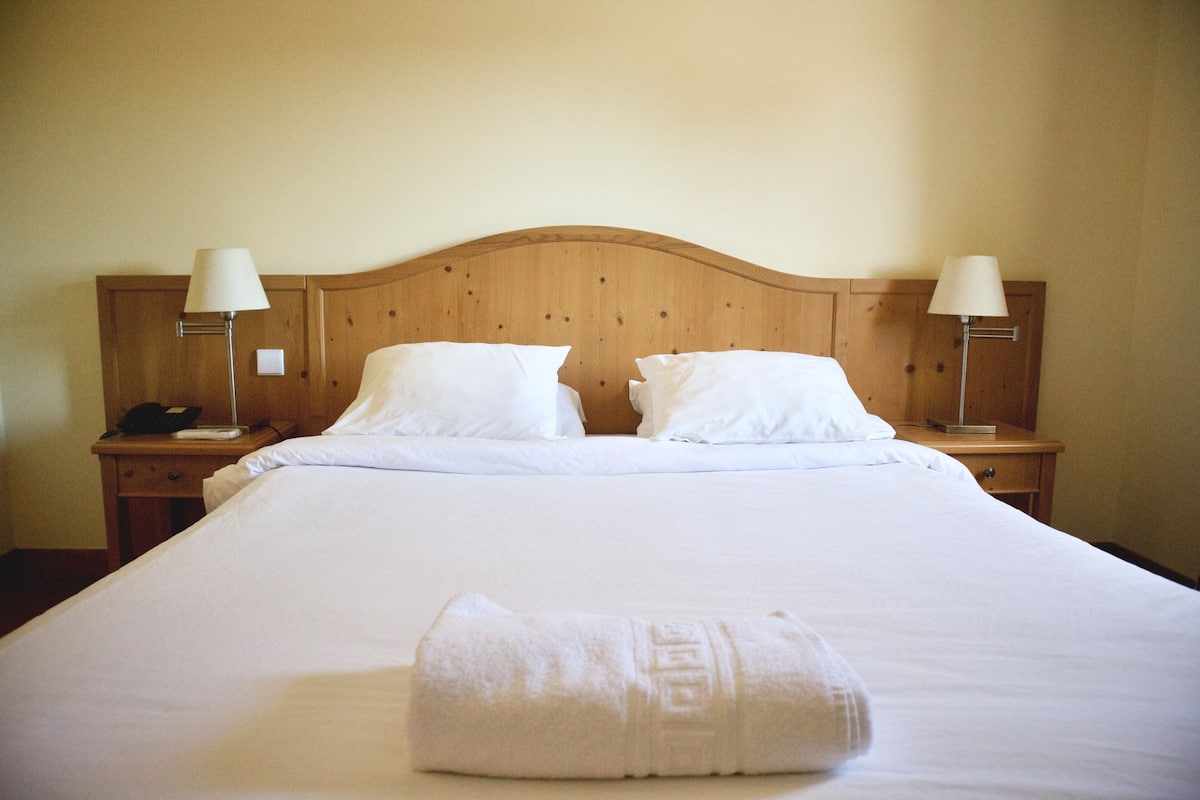 A neatly made bed with white linens and pillows, two bedside lamps, and a folded towel on the bed.
