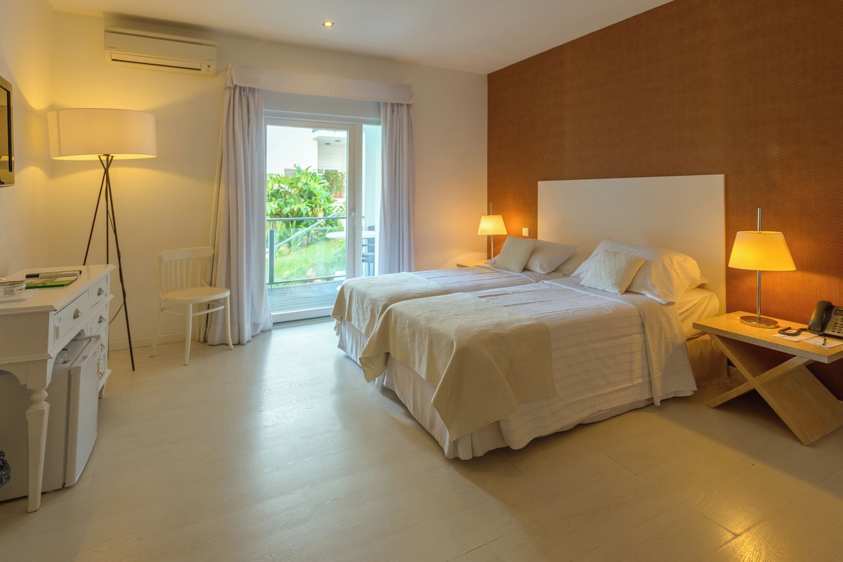 Habitación de hotel con dos camas, un escritorio con silla, una lámpara y vistas al verdor por la ventana.