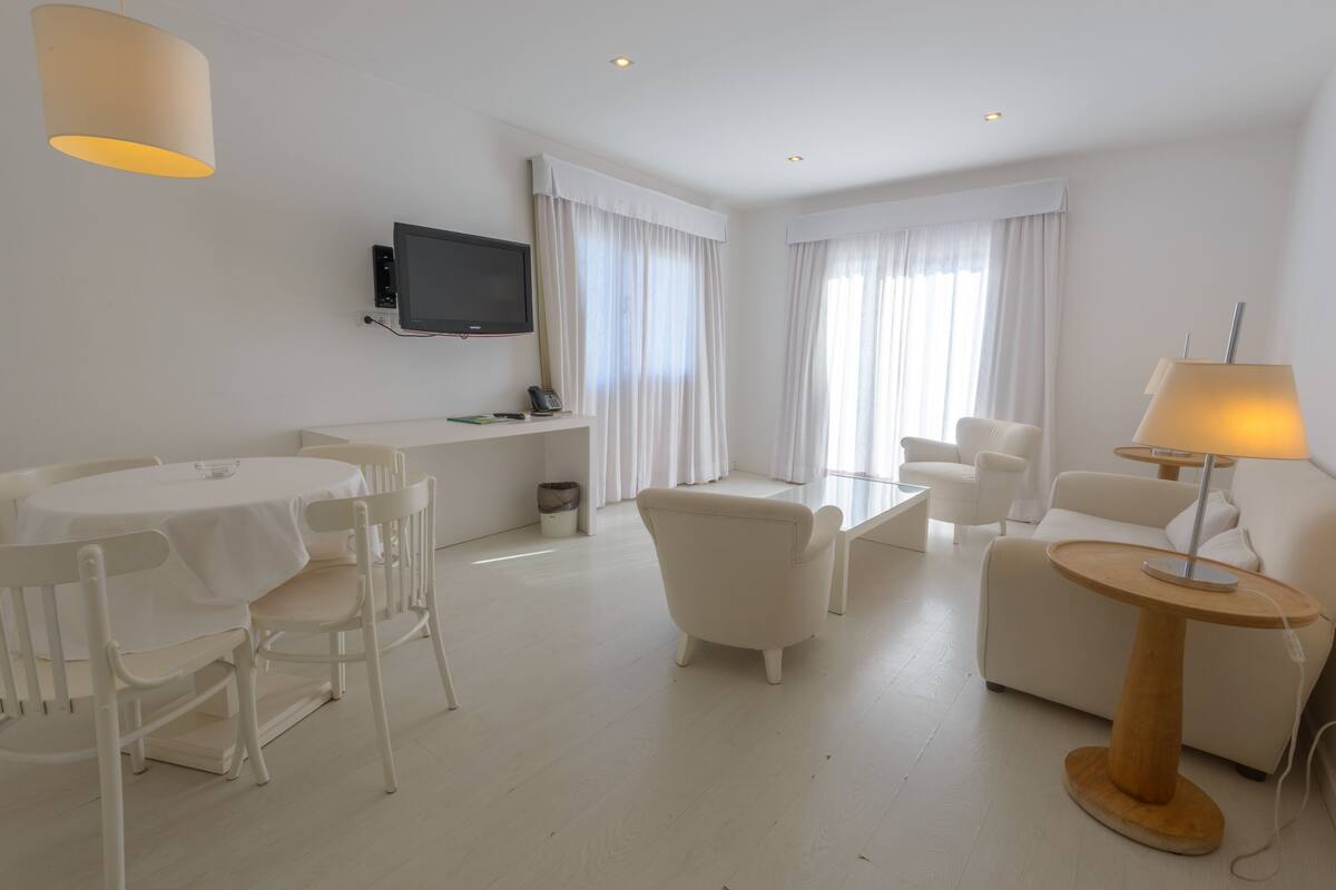 A modern, white-themed living room with a dining area, a sofa, a coffee table, and a TV mounted on the wall.