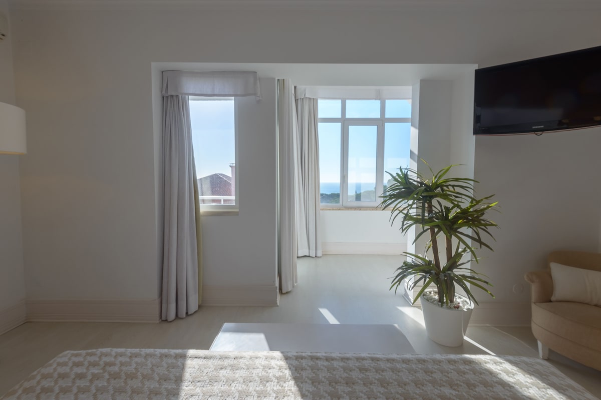 Ein geräumiges Zimmer mit großem Fenster, einem Blumentopf, einem Flachbildfernseher und einem beigefarbenen Sofa.