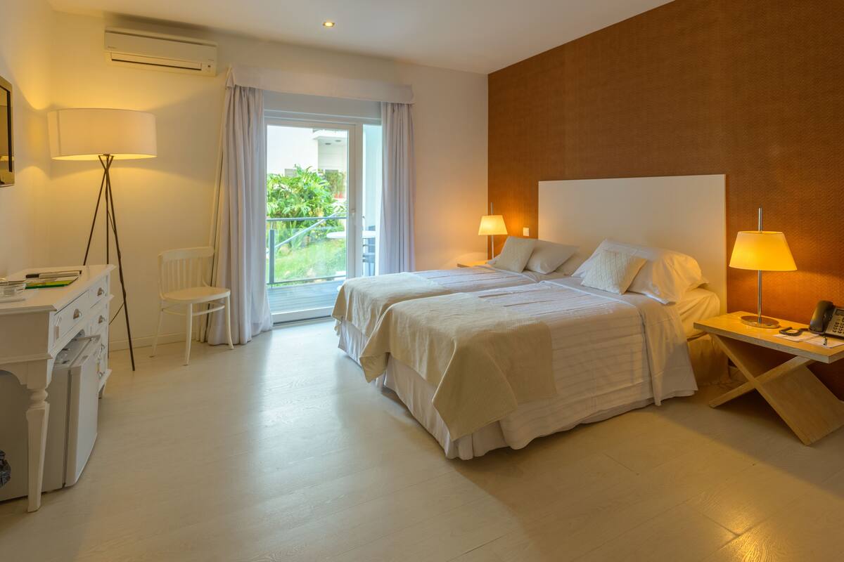 A hotel room with two beds, a desk with a chair, a lamp, and a view of greenery through the window.