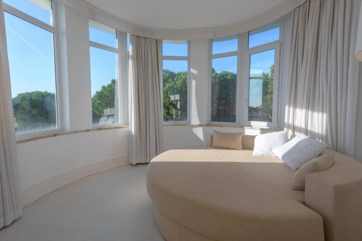 A modern bedroom with a curved bed, large windows, and a view of greenery.