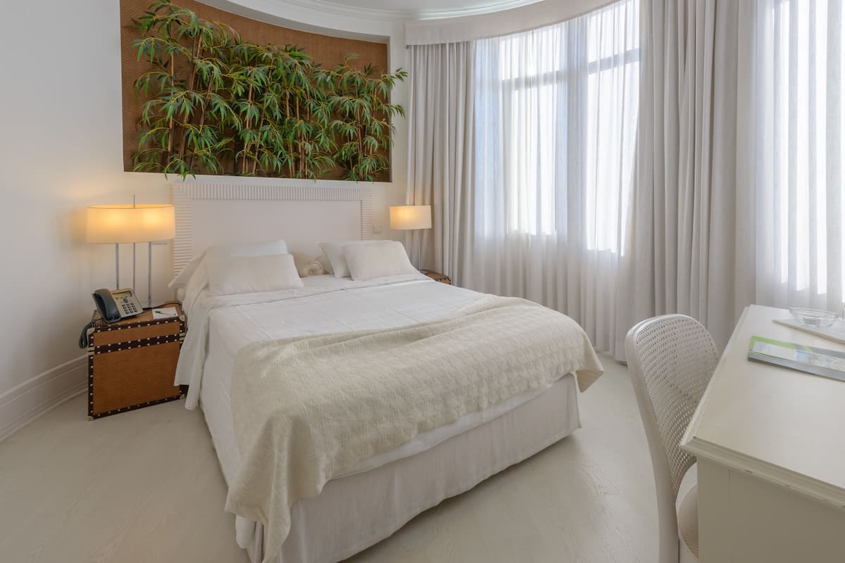 A hotel room with a large bed, two bedside lamps, a small table with a chair, a plant on the wall, and a window with curtains.