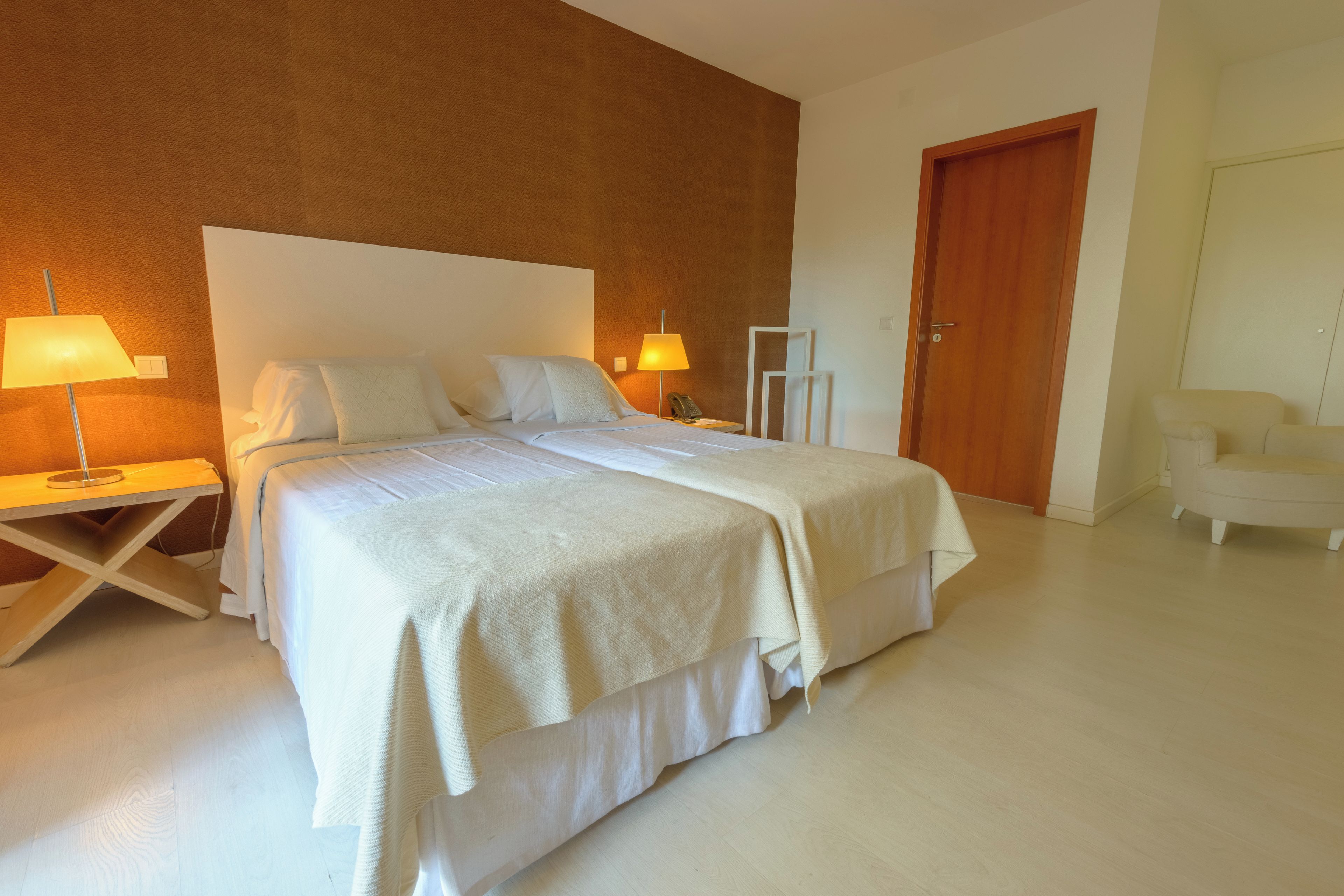 Uma cama bem arrumada com lençóis brancos e um cobertor bege, uma luminária de cabeceira e uma porta de madeira em um quarto de hotel moderno.