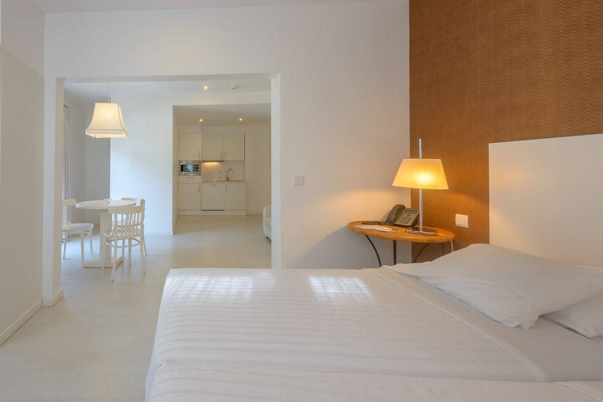 A hotel room with a bed, a desk with a lamp, and a view of the kitchen and dining area.