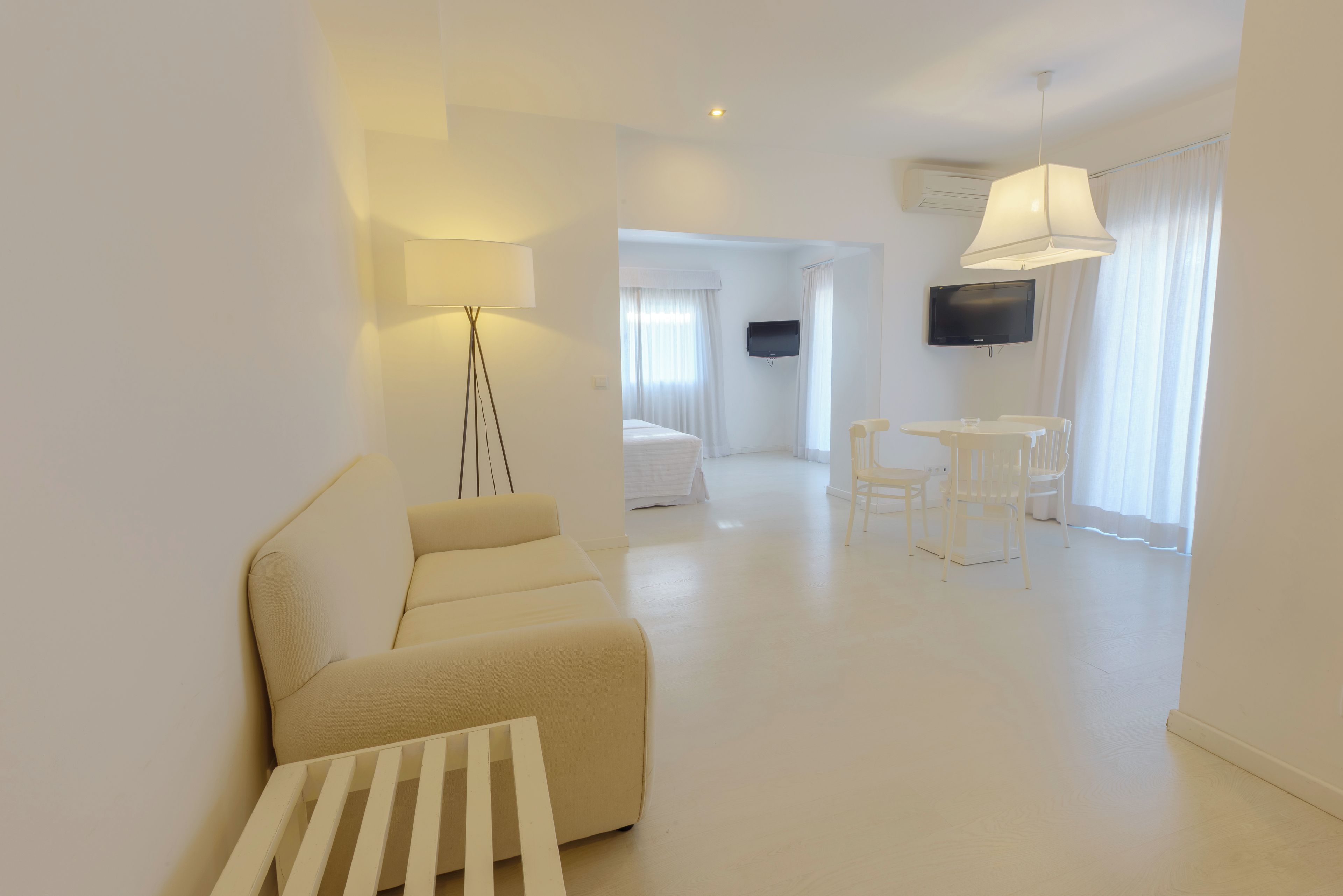 A modern, minimalist hotel room with a sofa, a small table, a TV, and a dining area with chairs.