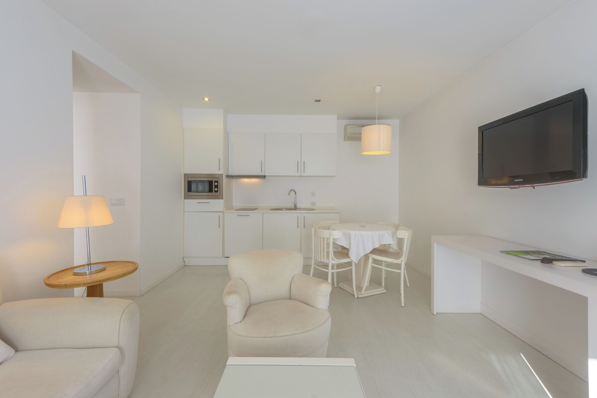 Una sala de estar moderna y minimalista con un sofá, una butaca, una mesita de noche con una lámpara, un comedor con mesa y sillas, una cocina con armarios y un microondas, y un televisor montado en la pared.