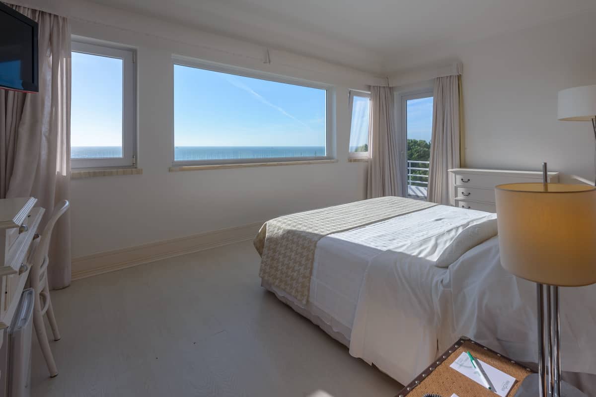 A hotel room with a bed, a desk, a chair, a lamp, and large windows offering a view of the sea.