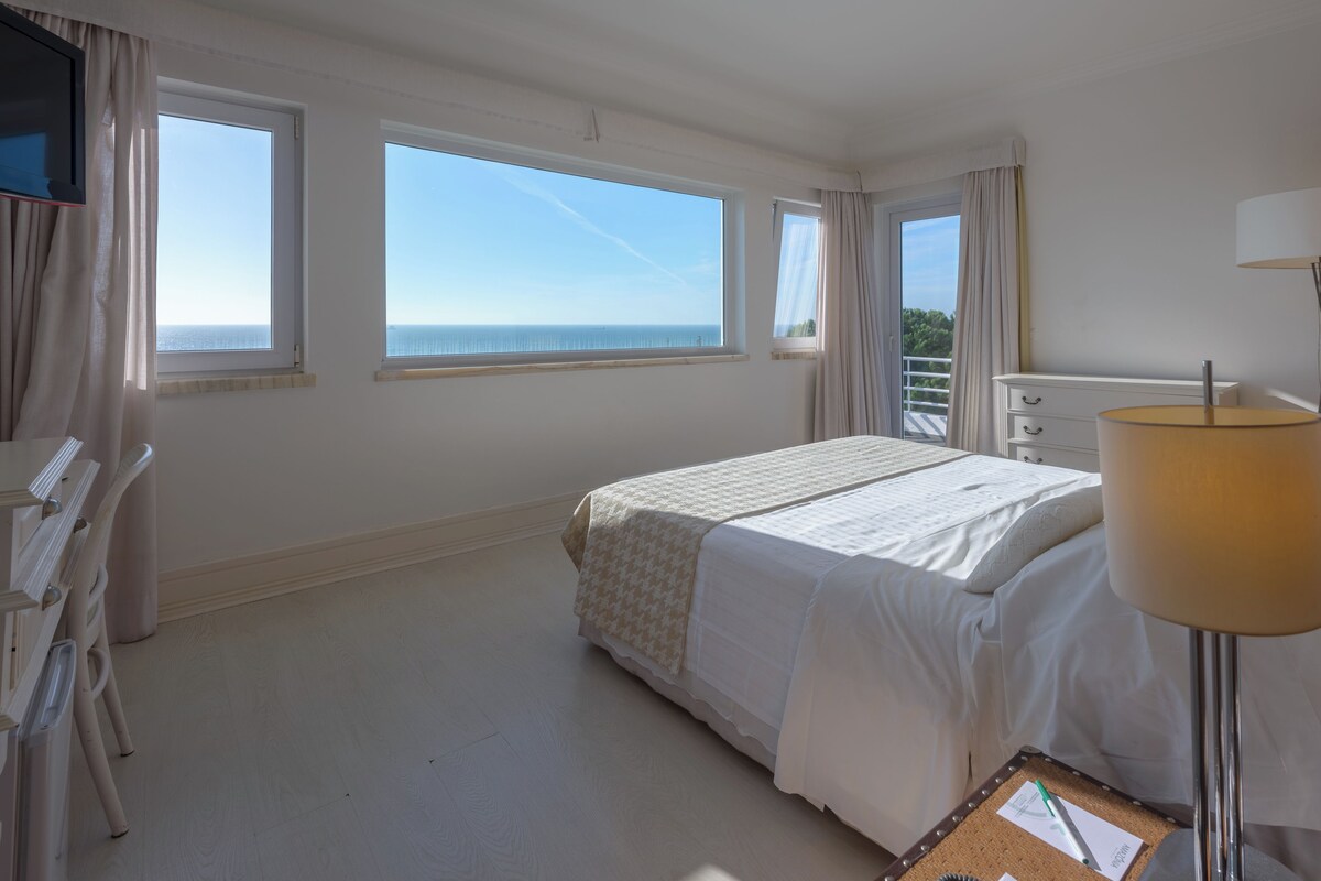 Habitación de hotel con cama, escritorio y silla. Dos ventanas ofrecen vista al mar.