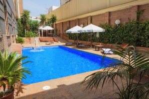 Ein Swimmingpool mit klarem, blauem Wasser, umgeben von Liegestühlen und Sonnenschirmen, mit einer Ziegelmauer und viel Grün im Hintergrund.