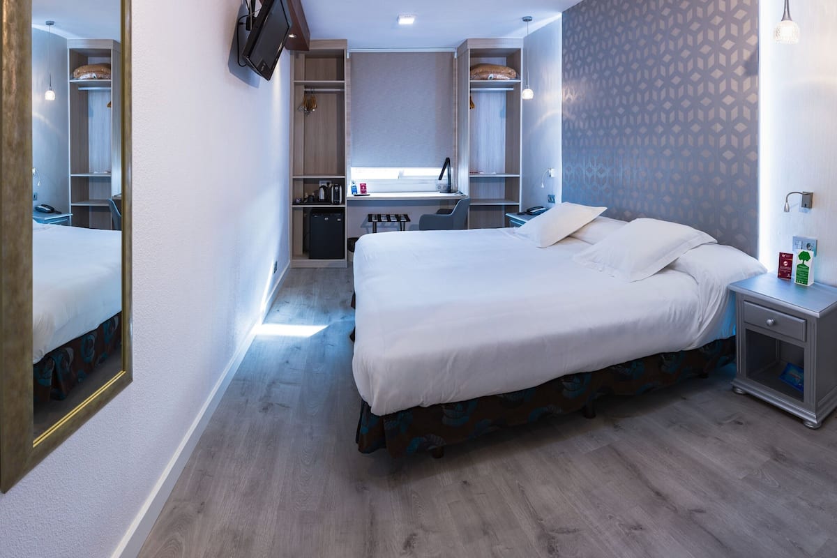 A hotel room with a bed, bedside table, mirror, wardrobe, desk, and a small kitchenette.