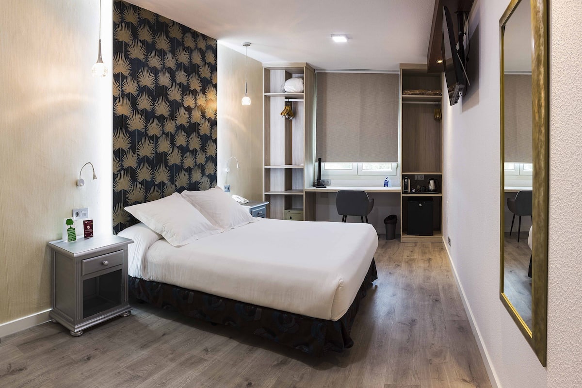 A hotel room with a bed, bedside table, lamp, desk, chair, wardrobe, and a mirror.