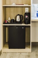 Una kitchenette compacta con un refrigerador negro, una cafetera, un hervidor y una máquina de café sobre un estante de madera.
