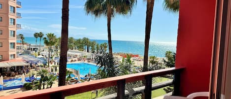 A balcony with a pool, palm trees, and a view of the beach and ocean.