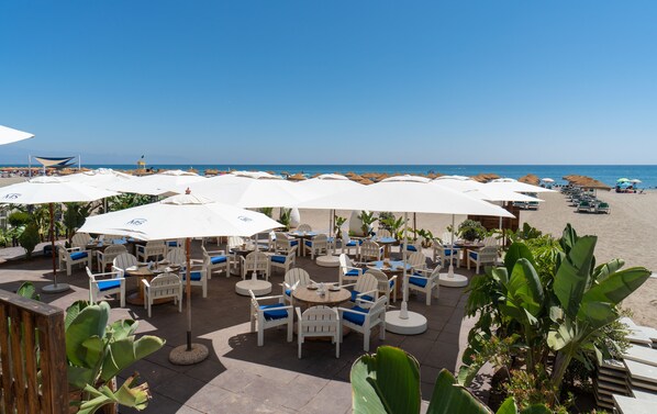 Ein Sitzbereich im Freien am Strand mit weißen Sonnenschirmen und Stühlen, mit Blick auf das Meer.