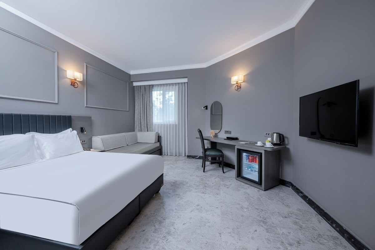 A modern hotel room with a large bed, a desk with a chair, a television, and a minibar.