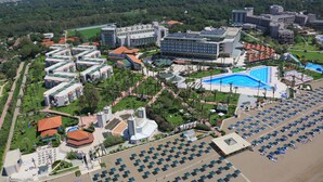 An aerial view of a resort with multiple buildings, a large swimming pool, and a parking area with numerous umbrellas. - ADORA HOTEL & RESORT - All inclusive (Serik)