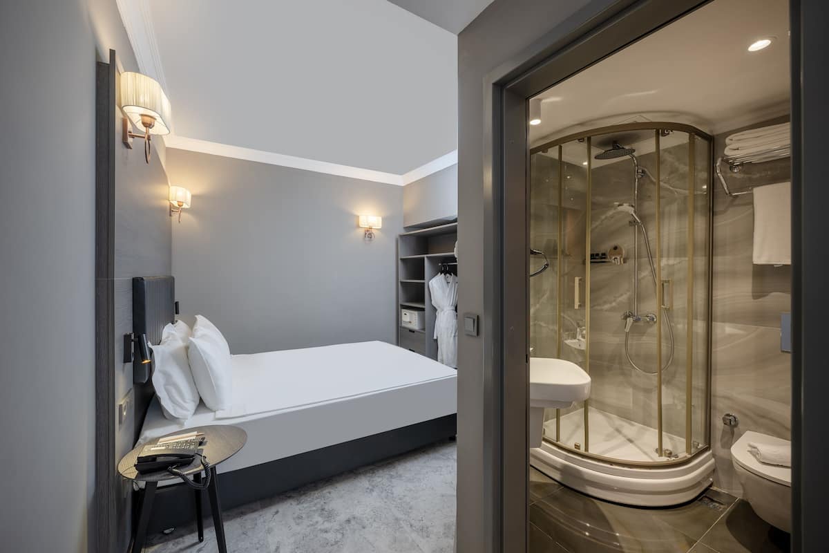 Ein modernes Hotelzimmer mit Bett, Nachttisch, Lampe und einer Duschkabine aus Glas.