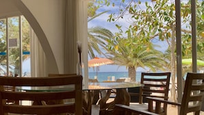 Une pièce dotée d’une grande fenêtre donnant sur une plage, de chaises en bois, d’une table en verre et offrant une vue sur les palmiers et la mer.