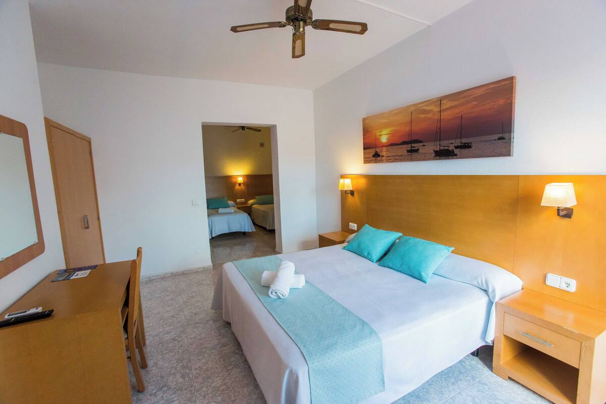 Een hotelkamer met een bed, een bureau, een stoel, een plafondventilator, een schilderij van boten aan de muur, en een nachtkastje met een lamp.