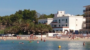 Een strand met mensen die zwemmen en zonnebaden, palmbomen en een wit gebouw met balkons.