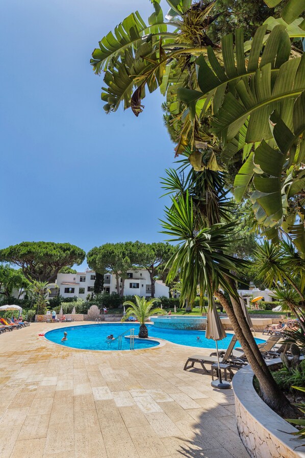 A pool area with lounge chairs and umbrellas, surrounded by tropical plants and a clear blue sky.