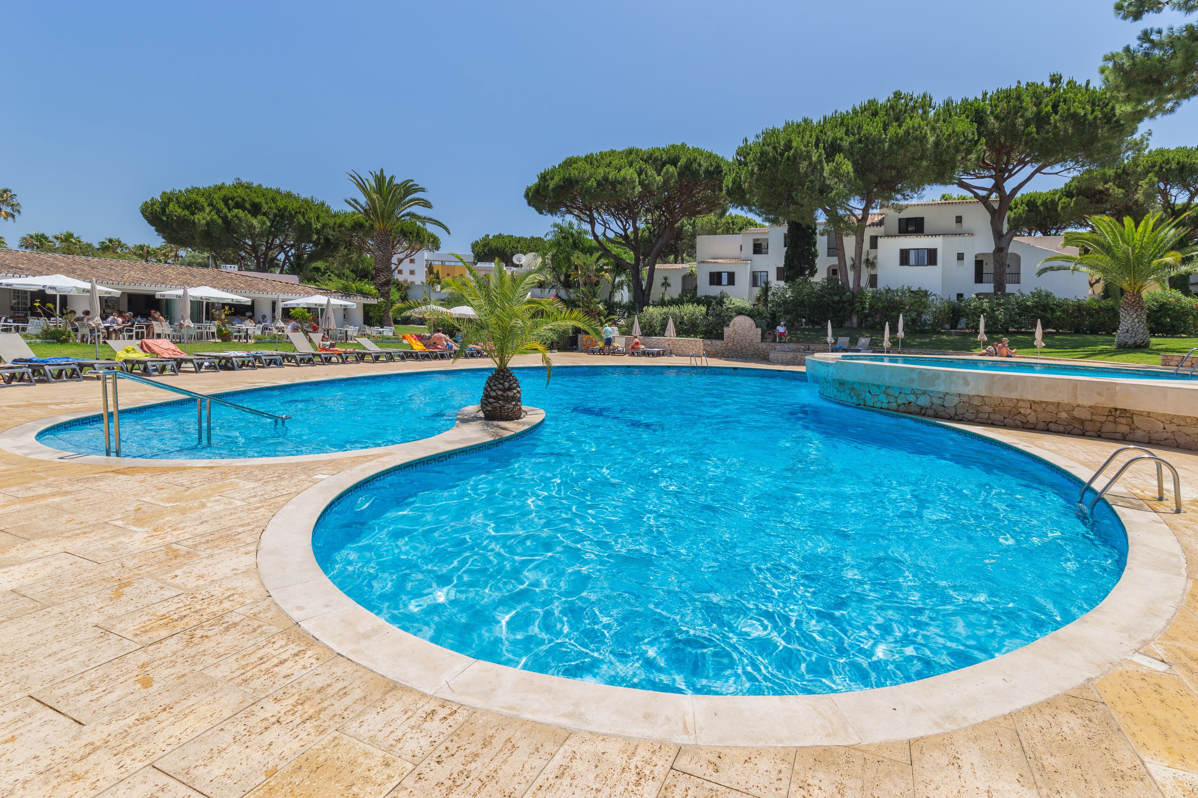 A swimming pool with lounge chairs and umbrellas, surrounded by trees and buildings.