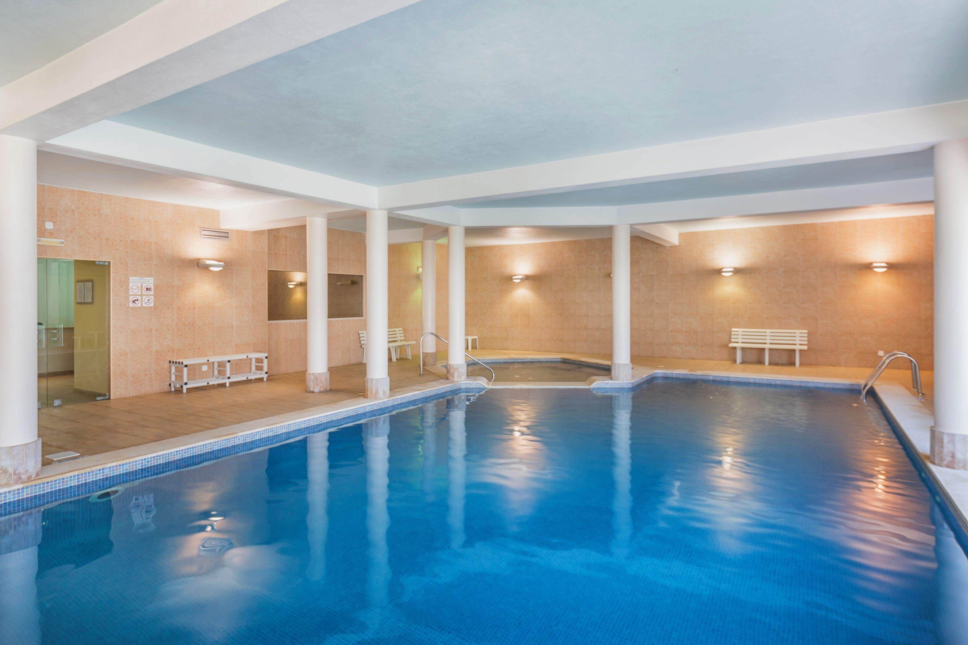 Una piscina interior con agua cristalina, columnas blancas y paredes de azulejos.