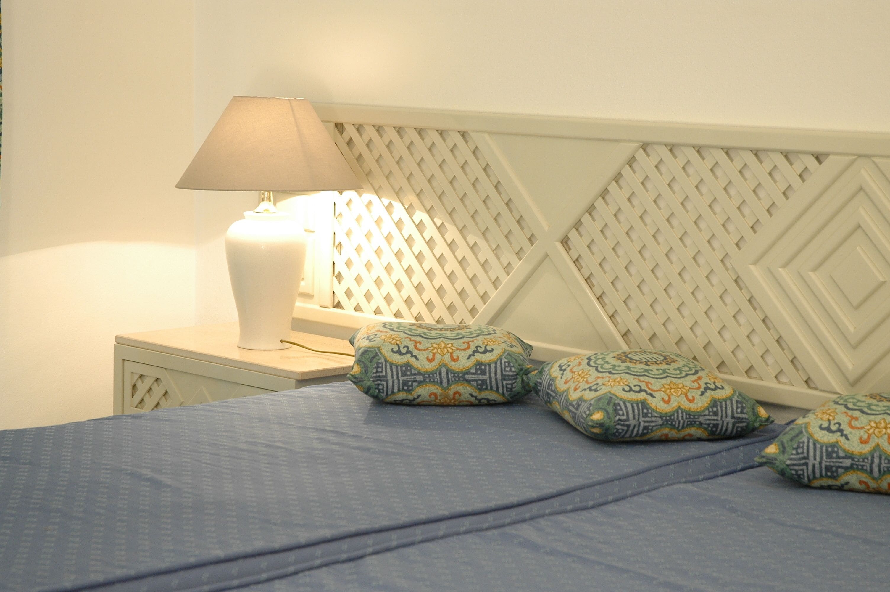 A neatly made bed with a blue bedspread and decorative pillows, a bedside lamp, and a patterned headboard.
