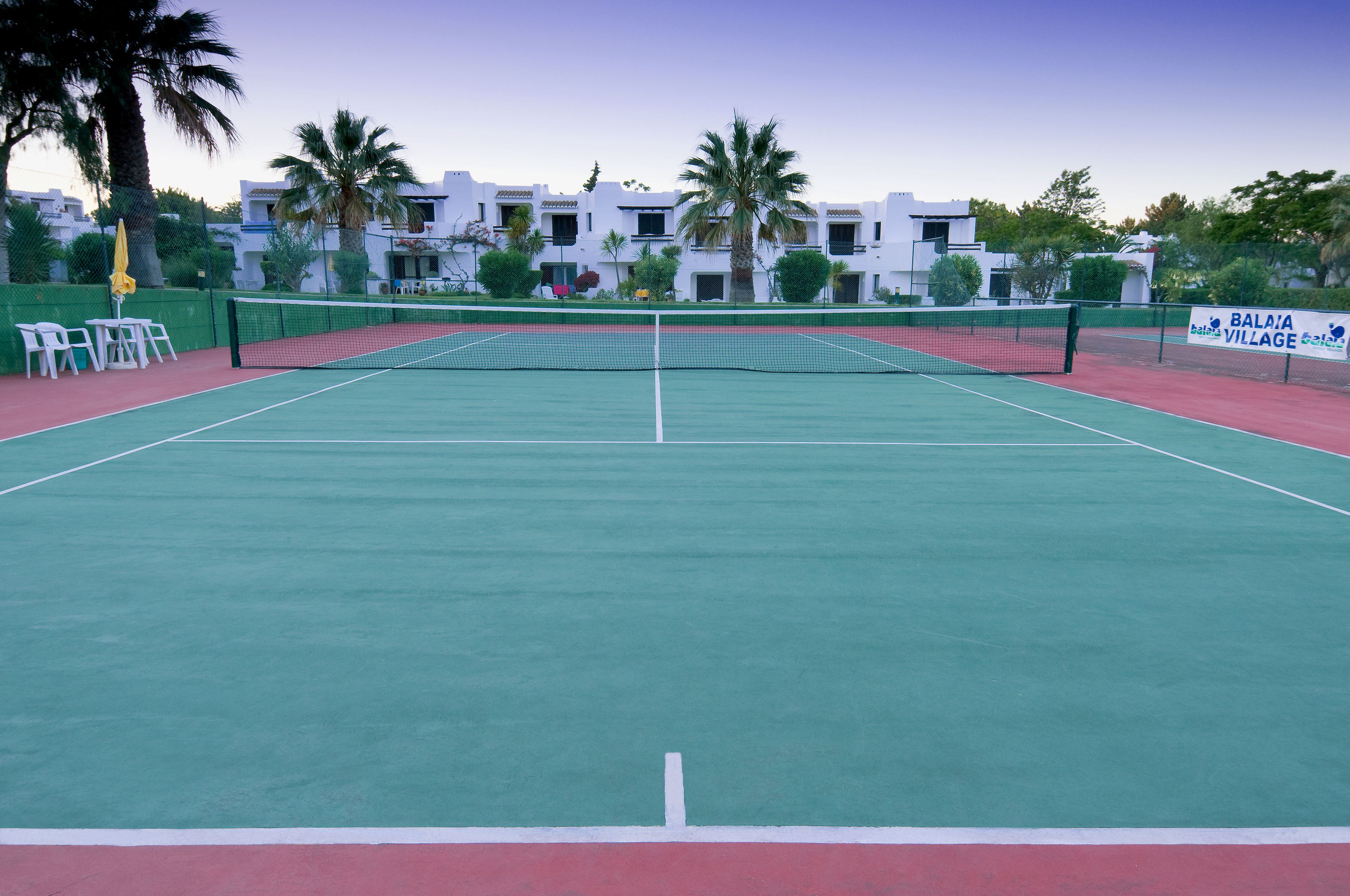 Een tennisbaan met een net, omgeven door een groene hekken, en een bordje met BALAIA VILLAGE erop.