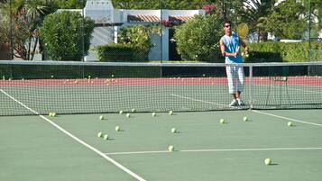 A tennis court with a net in the middle, surrounded by greenery and a building in the background.