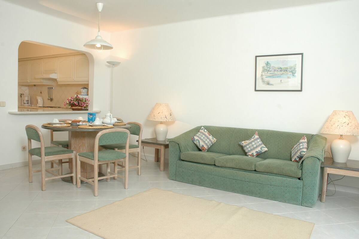 A living room with a green sofa, two side tables with lamps, a dining table with chairs, and a kitchen area visible through an archway.