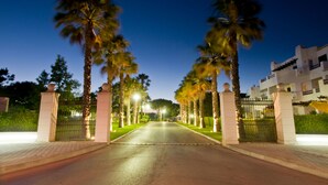 A gated entrance with palm trees, a paved road, and a building with balconies in the background. - Balaia Golf Village Resort (Albufeira)