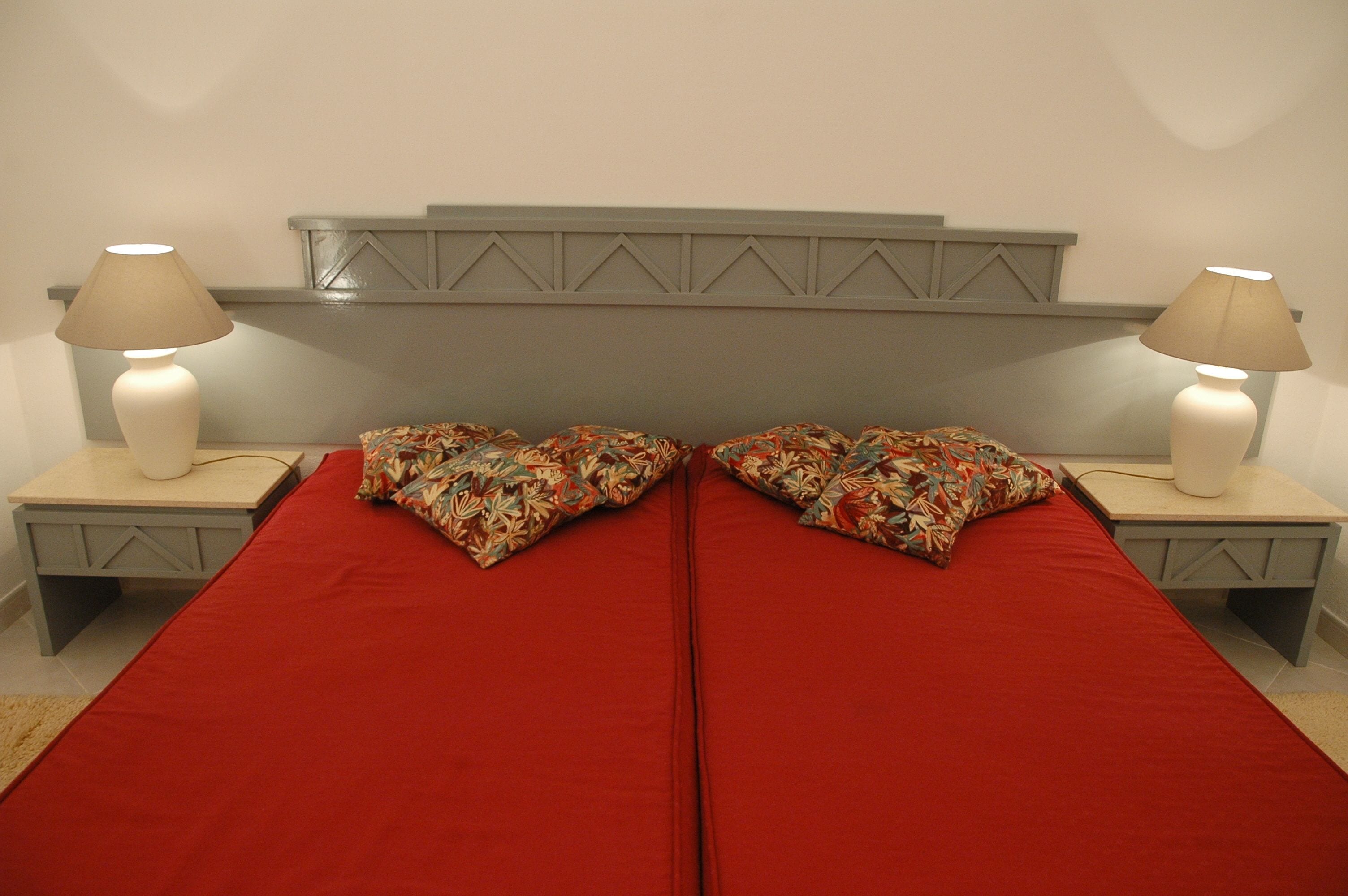 Ein Doppelbett mit roter Bettwäsche und Kissen mit Blumenmuster, flankiert von zwei Nachttischen mit Lampen, an einer schlichten Wand mit einem dekorativen Kopfteil.