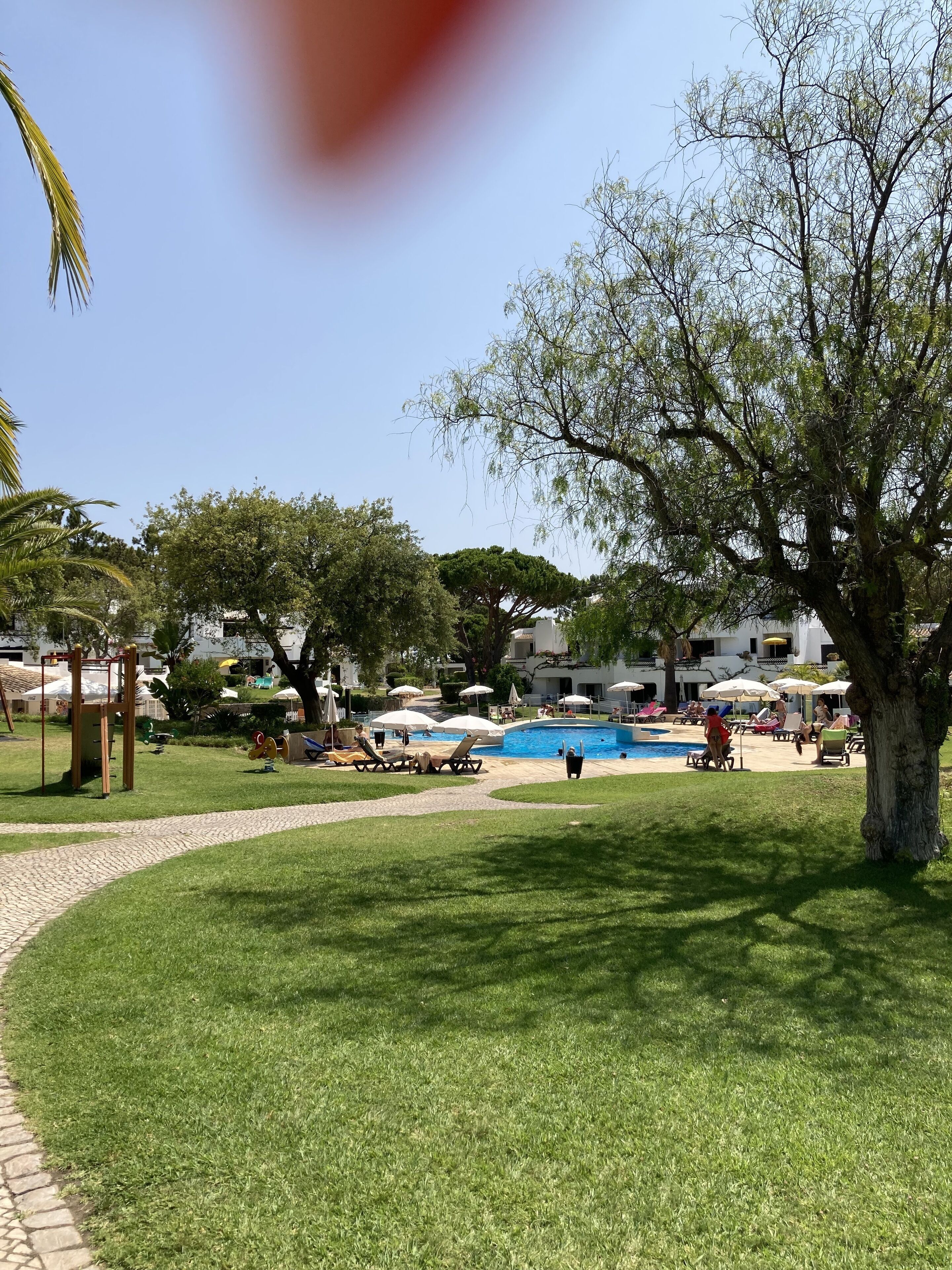A sunny outdoor area with a swimming pool, lounge chairs, and a playground.
