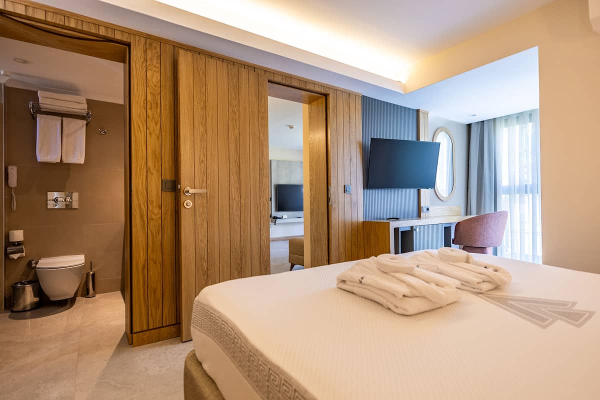 A hotel room with a large bed, a wooden wardrobe, a bathroom with a toilet and sink, a desk with a chair, and a flat-screen TV mounted on the wall.